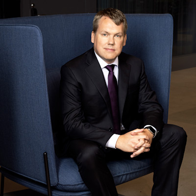 Stefan Widing wearing a dark suite and a purple tie, sitting in a modern, dark blue armchair